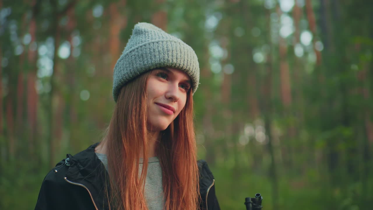 Close up of woman in knit hat holding pump nozzle while standing in forest clearing, softly gazing toward someone in background with slight expression, surrounded by blurred green trees