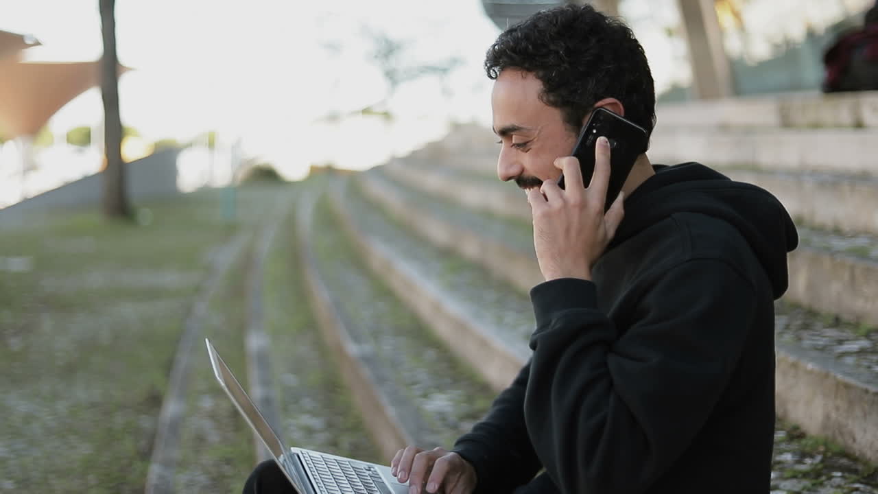 vista lateral de un hombre árabe con cabello rizado oscuro y barba con capucha negra sentado en las escaleras afuera