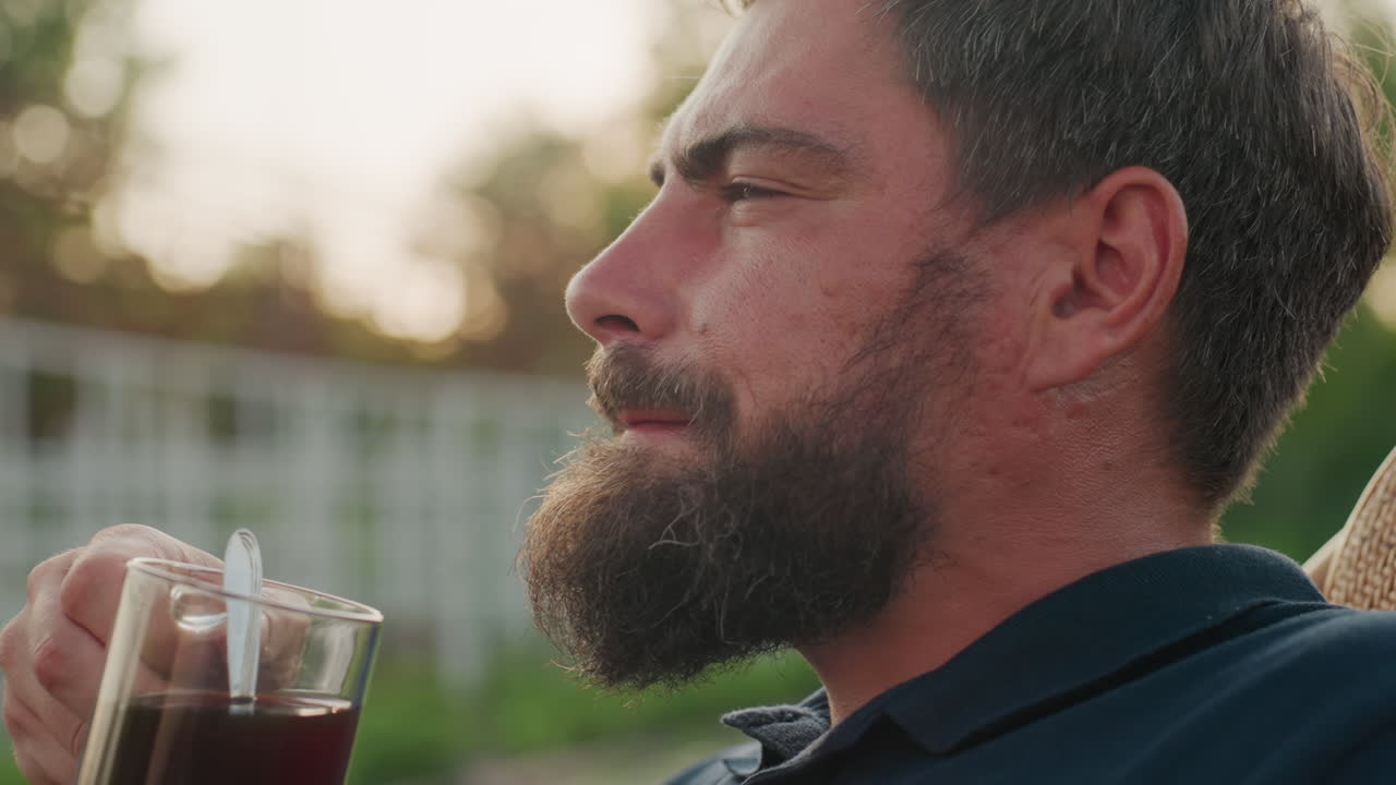 close up side view man sipping hot black coffee from clear glass mug seated in wicker rocker in garden at golden hour sunlight creating warm soft glow and bokeh foliage background