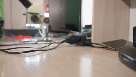 a close-up view of a hand wearing a black dropping glasses on a wooden desk in a lab setting, with electronical tools on the table and wires around