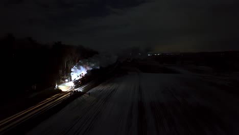 a train moves along snowy tracks during nighttime, releasing smoke into the air. the surrounding landscape is quiet and dark, punctuated by distant lights and a small station.