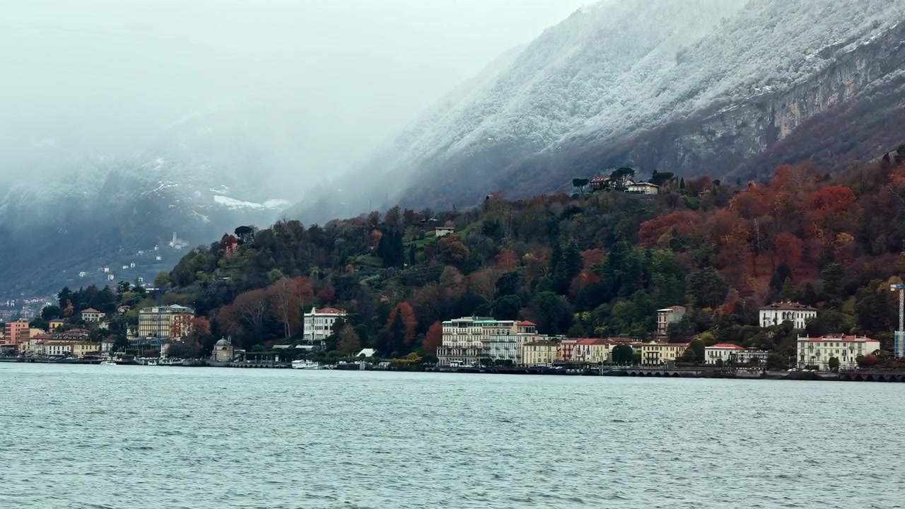 Carrellata della telecamera verso destra sulle acque del Lago di Como che rivela il lungolago di Bellagio con il fogliame autunnale e le montagne innevate, ritraendo la bellezza drammatica dei laghi italiani nel tardo autunno.