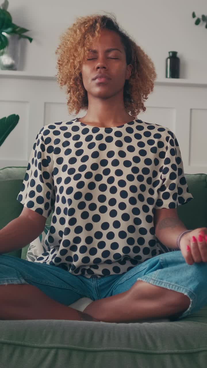 Young beautiful african american woman meditates sits on sofa in lotus position