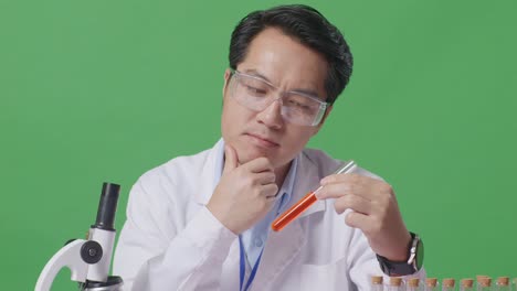 close up of asian man scientist looking at test tube then thinking, looking around, and raising index finger while working on the table with microscope in the green screen background laboratory