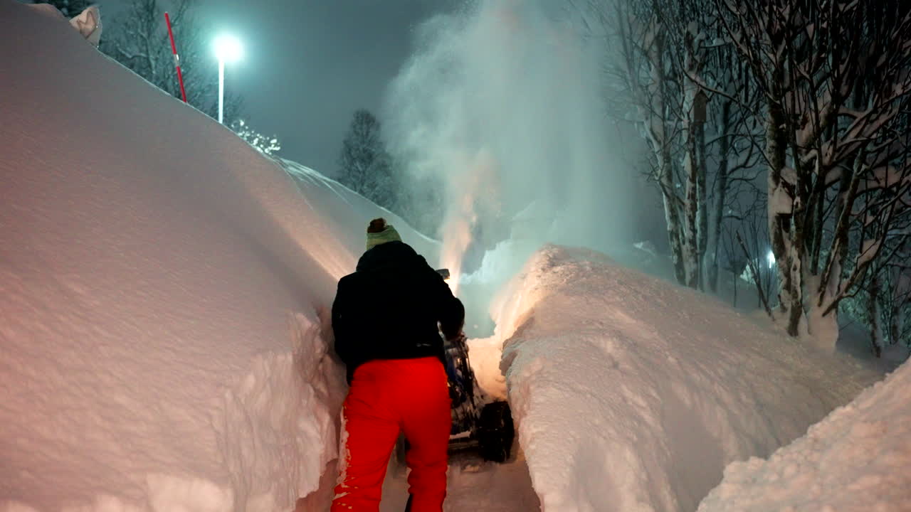 A snowplow clearing heavy snow in Tromsø, Norway, urban winter night along snowy path, rearview of person in orange pants