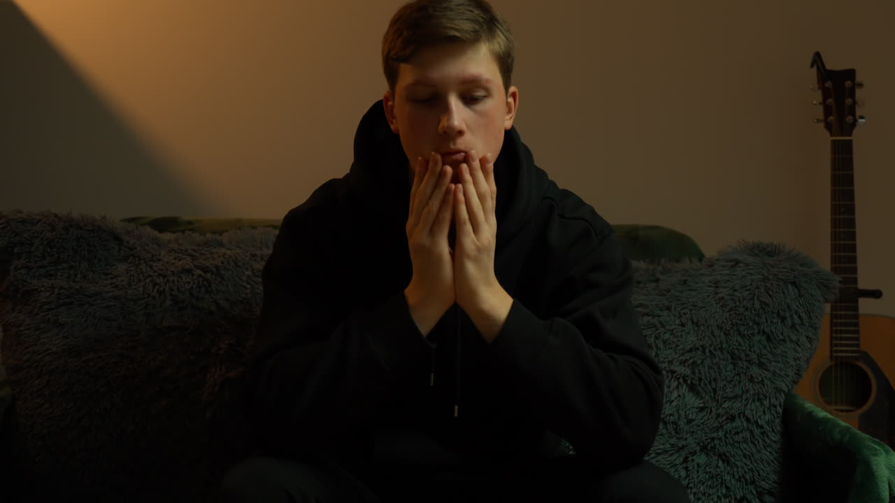 Young caucasian man in his 20s showing contemplation and deep thought, hands clasped together, looking concerned and pensive in dim room at home, feeling stressed with acoustic guitar in background