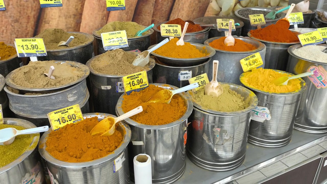 A variety of colorful spices are neatly displayed for sale in clean stainless steel jars. Each container features a clear price tag. The organized market setup looks hygienic and appealing.