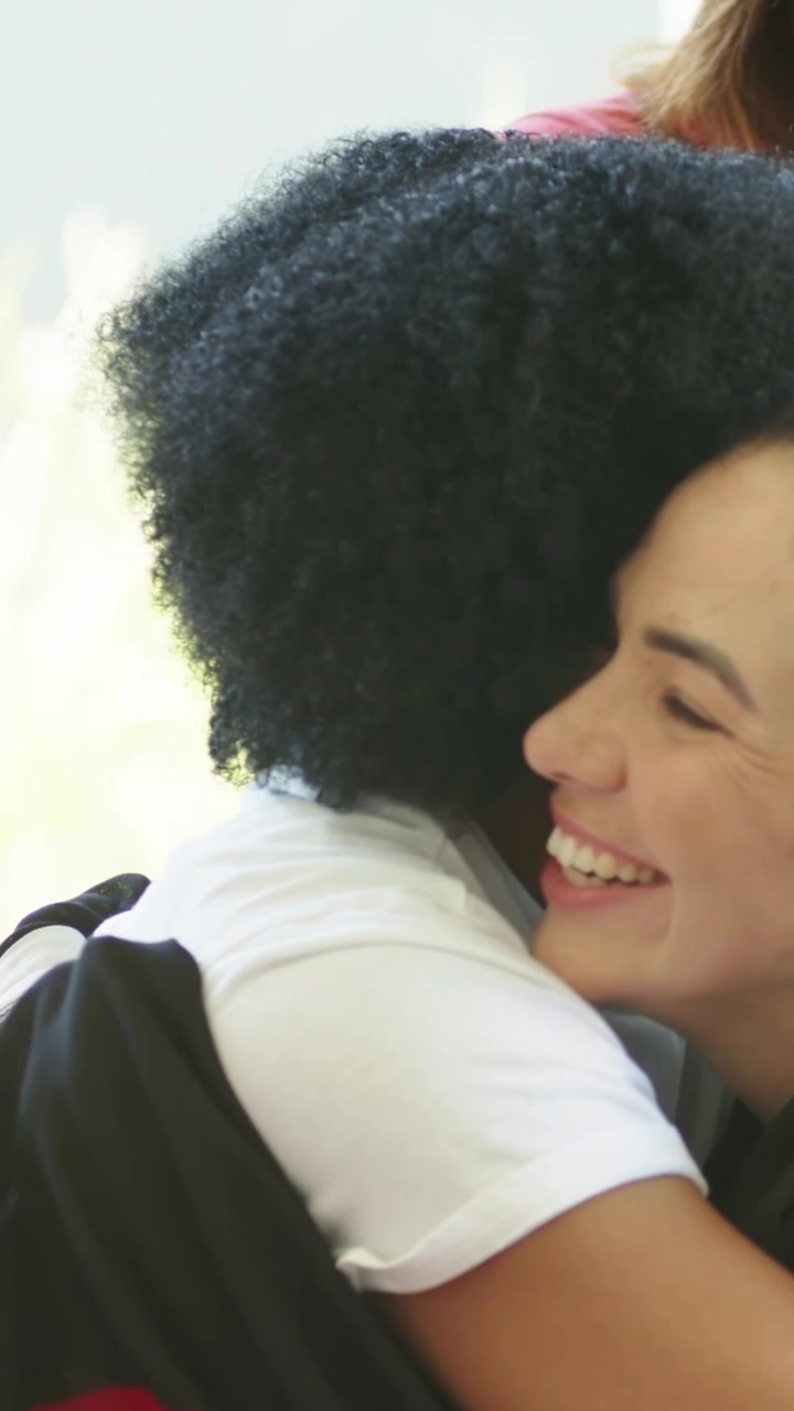 Video verticale: Donna riccia appoggiata che saluta un'amica con un abbraccio vicino alla finestra, maglietta bianca, spallina dello zaino
