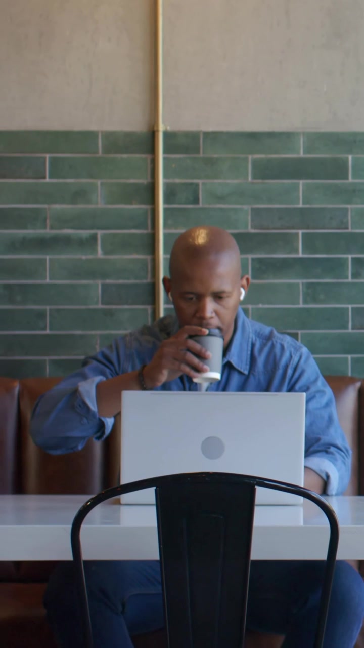 Vertical video: Looking at laptop, man in blue shirt typing and sipping coffee cup for work at cafe