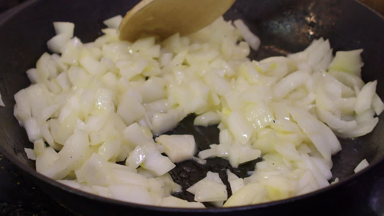Close up: Wooden spoon stirs diced onions frying in hot cast iron pan