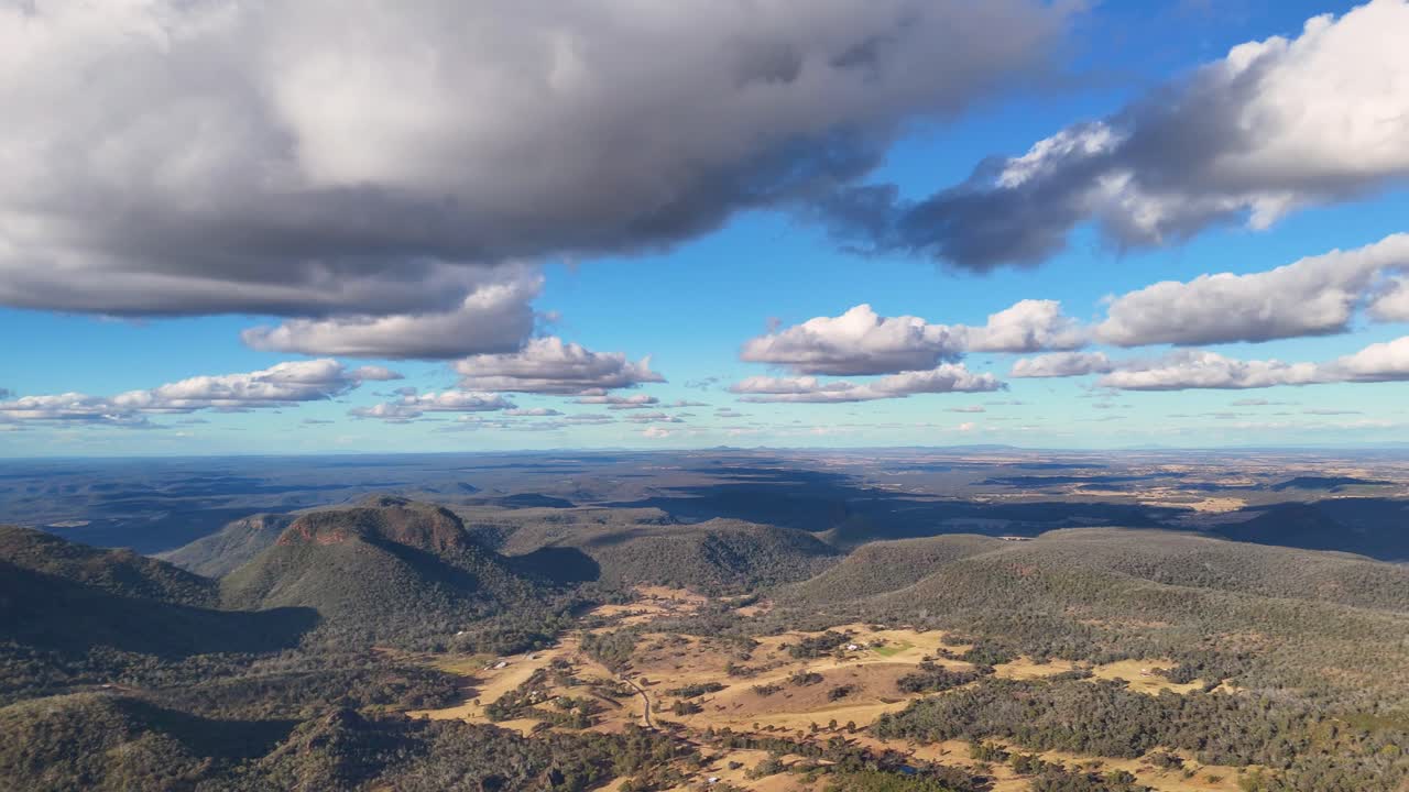 Drone captures sweeping panoramic view of rugged bushland, gum trees, and dramatic clouds over Warrumbungle National Park in afternoon sunlight, with smooth lateral camera movement