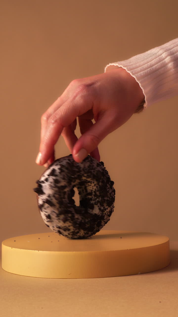 Close up of hand spinning chocolate donut with white icing and dark cookie crumbs on round pedestal. Warm beige studio background with clean minimal aesthetic and soft light