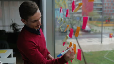 smiling handsome young man writing on sticky notes on window.