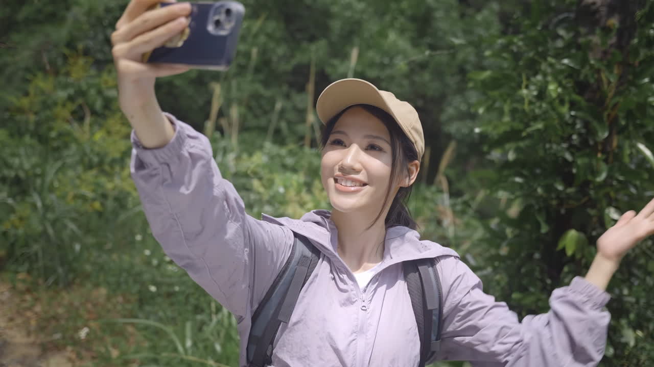 Mulher sorridente, caminhante e mochileira, alcançando o topo de um pico de montanha, descansando, fazendo uma videochamada transmitindo ao vivo no smartphone com um aceno, sorriso ou contato durante a pausa para mostrar a bela paisagem na mata.
