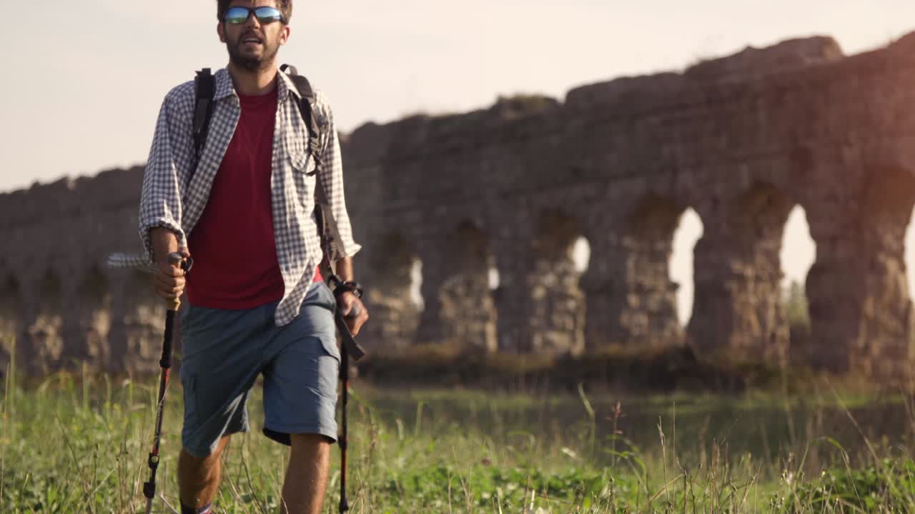 젊은 배 타는 남자가 황혼이 지는 로마의 파르코 델리 아키도티 공원 유적에 있는 로마 수로 앞에서 잔디 위에 지이를 들고 맨발로 고 있다.