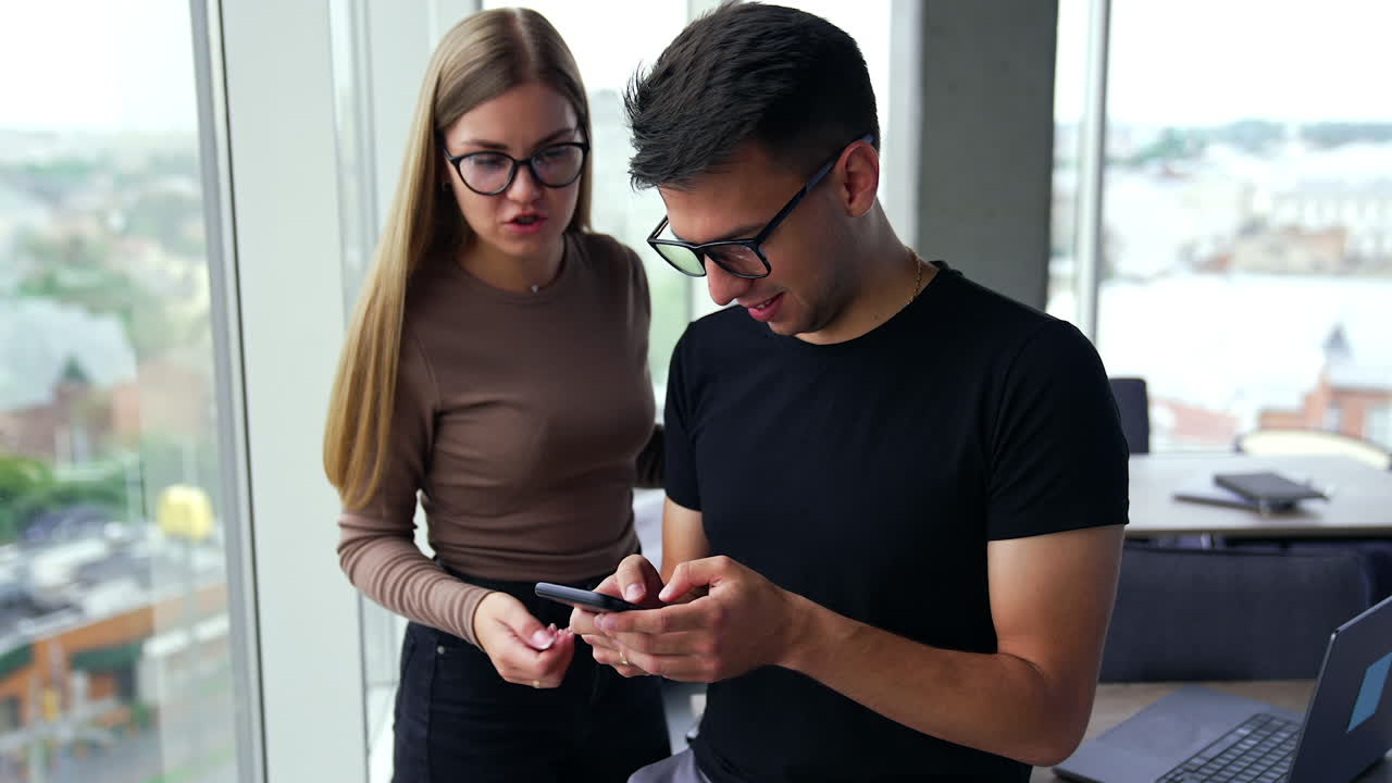 Young diverse employees in office stand at the window looking at phone. Lady telling something to a man typing on the smartphone. Blurred backdrop.