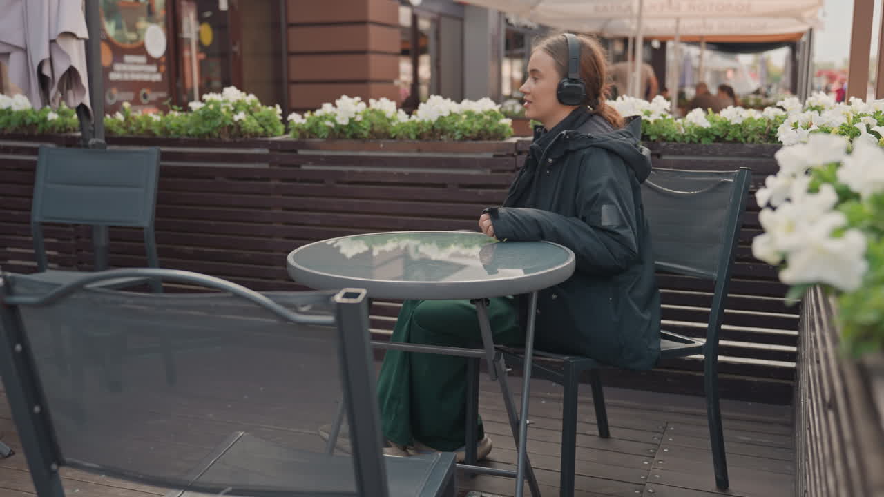 Una persona seduta al tavolino di un bar all'aperto si avvicina, si siede, si appoggia allo schienale e ascolta le cuffie mentre tiene in mano un bicchiere da asporto, indossa un cappotto scuro e una giacca, terrazza di un bistrot cittadino incorniciata da fioriere