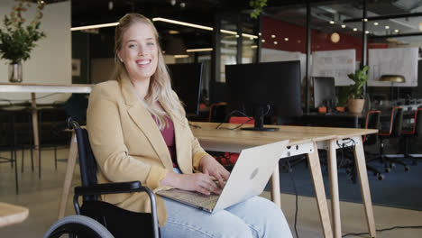 Portrait-of-happy-caucasian-businesswoman-in-wheelchair-using-laptop-in-office,-in-slow-motion