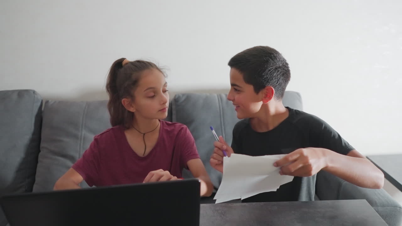 Un chico muestra alegremente su trabajo escrito a mano a una chica sentada a su lado, que sonríe mientras mira su papel y luego le da una palmada juguetona en la cabeza, haciéndolo reír y seguir escribiendo mostrando hoyuelos.