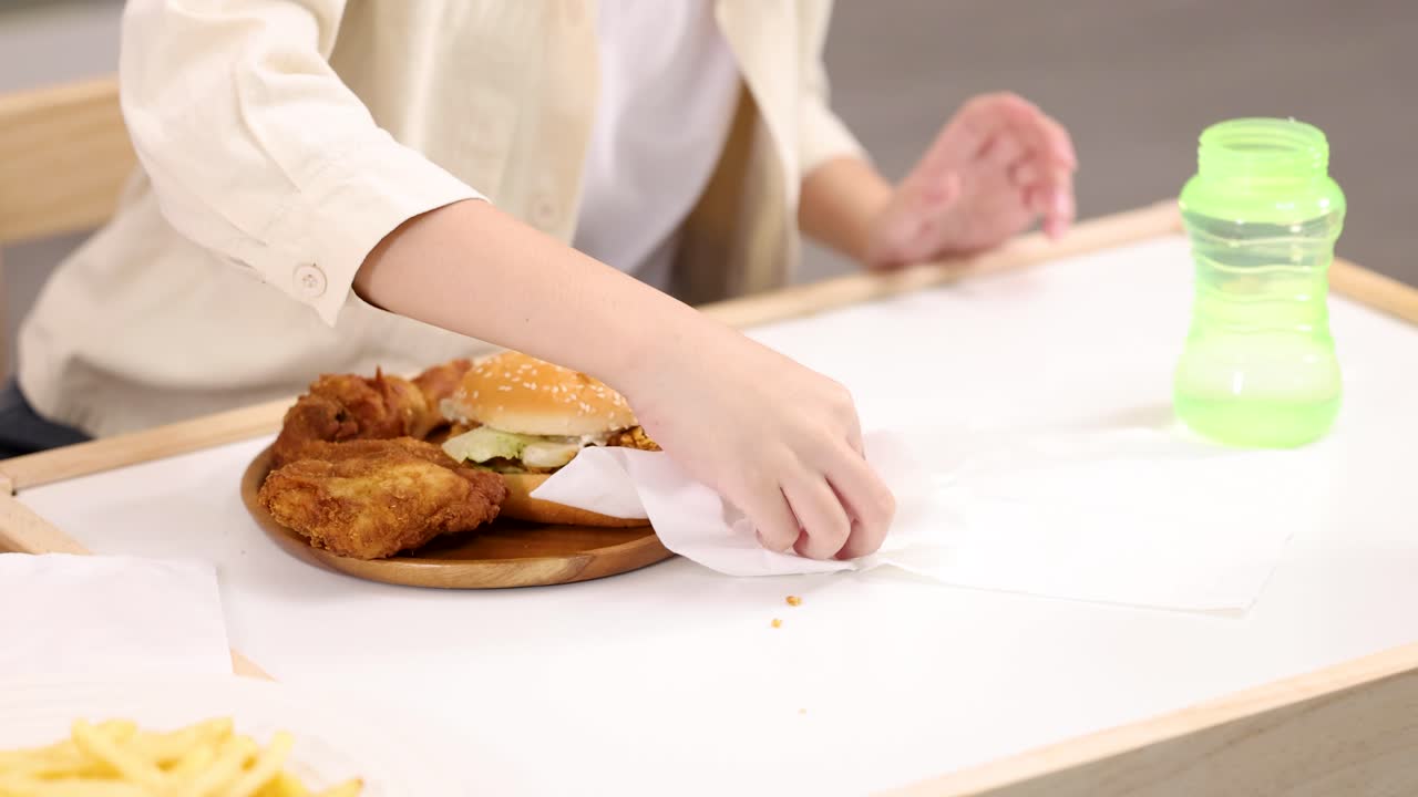Child wipes crumbs from white cafeteria table around fried chicken burger meal, static camera