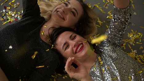 pretty young women lying on the floor full of golden confetti after a glamorous new years eve party, laughing and dancing