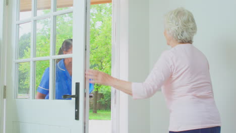 mujer anciana en casa usando un marco para caminar saludando a una enfermera o trabajadora de atención en uniforme en la puerta
