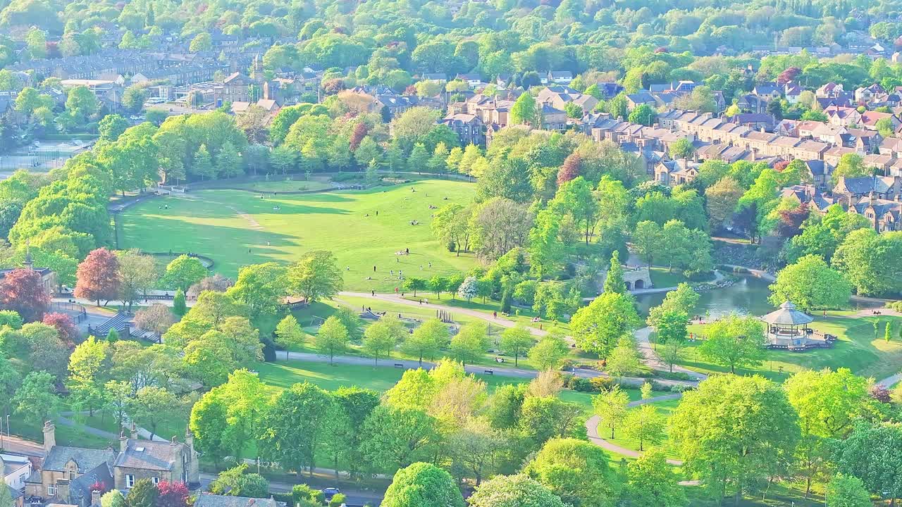 Aerial shot overlooking a green open parkland in Huddersfield near Springwood, with walking paths, scattered trees, and adjacent residential zones under soft golden light, aerial view, real time