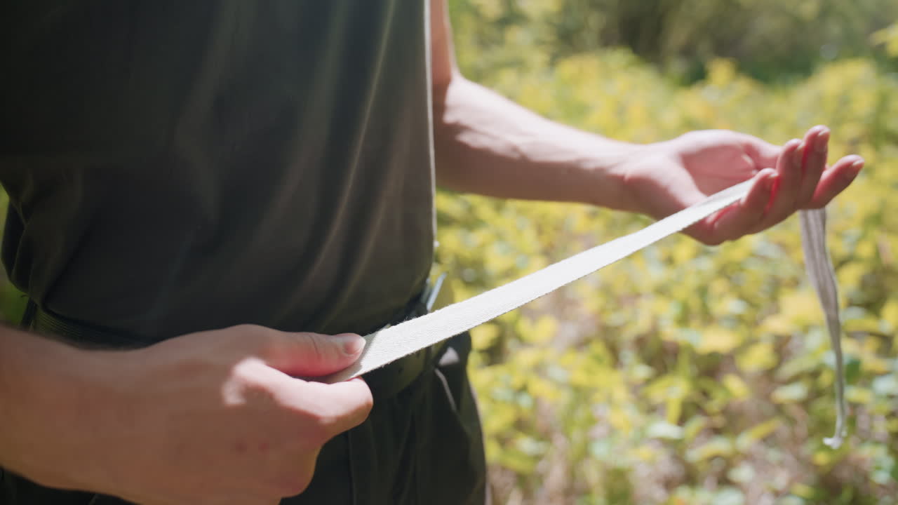 Mid shot of tourist holding white fabric headband, stretching and rolling it between palms over black belt and olive tank top, sunlight filtering through canopy of green foliage, veins on forearms