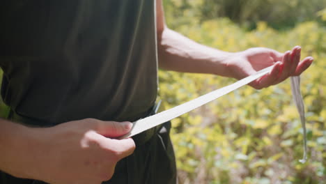 mid shot of tourist holding white fabric headband, stretching and rolling it between palms over black belt and olive tank top, sunlight filtering through canopy of green foliage, veins on forearms