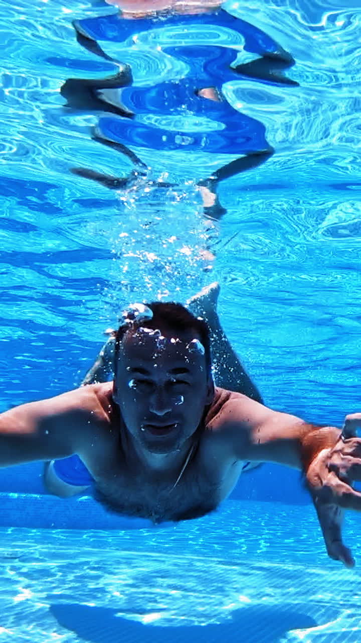 Vacation concept. Happy young man swimming inside the swimming pool. Man is diving under the water and moving forward to camera. Vertical video