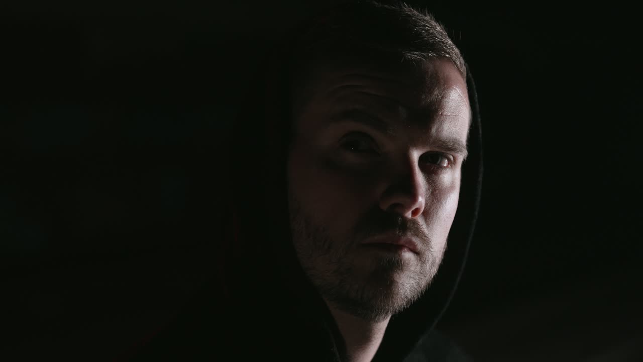 A man in the dark looks into the camera. his face is half-lit. he shows various emotions