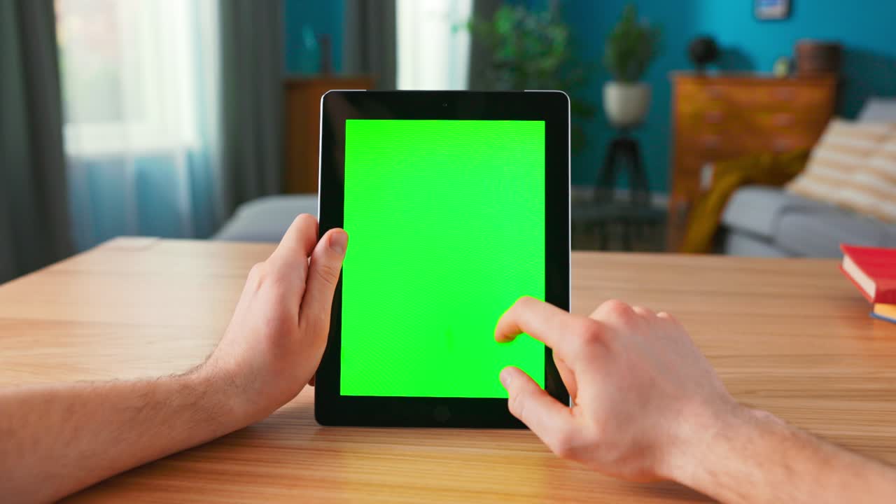 Close-up of Man Using Green Mock-up Screen Digital Tablet Computer in Portrait Mode while Sitting at His Desk. In the Background Cozy Living Room