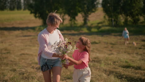 daughter gifting her mother beautiful flower bouquet with a warm smile in sunny farmland, blur view of another woman and dog in distance, with background of lush green trees and vast landscape