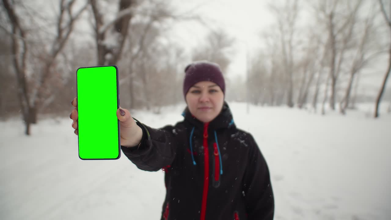 Woman shows a phone with chromakey in winter the park. Woman stands under the snowfall and holds a phone with green screen in hand