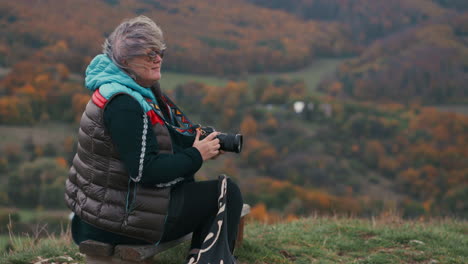 Female-Photographer-sitting-on-a-bench-taking-pictures-with-her-camera-and-looking-around-while-wind-is-blowing-up-her-grey-hair,-surrounded-by-beautiful-autumn-nature-with-colourful-orange-trees