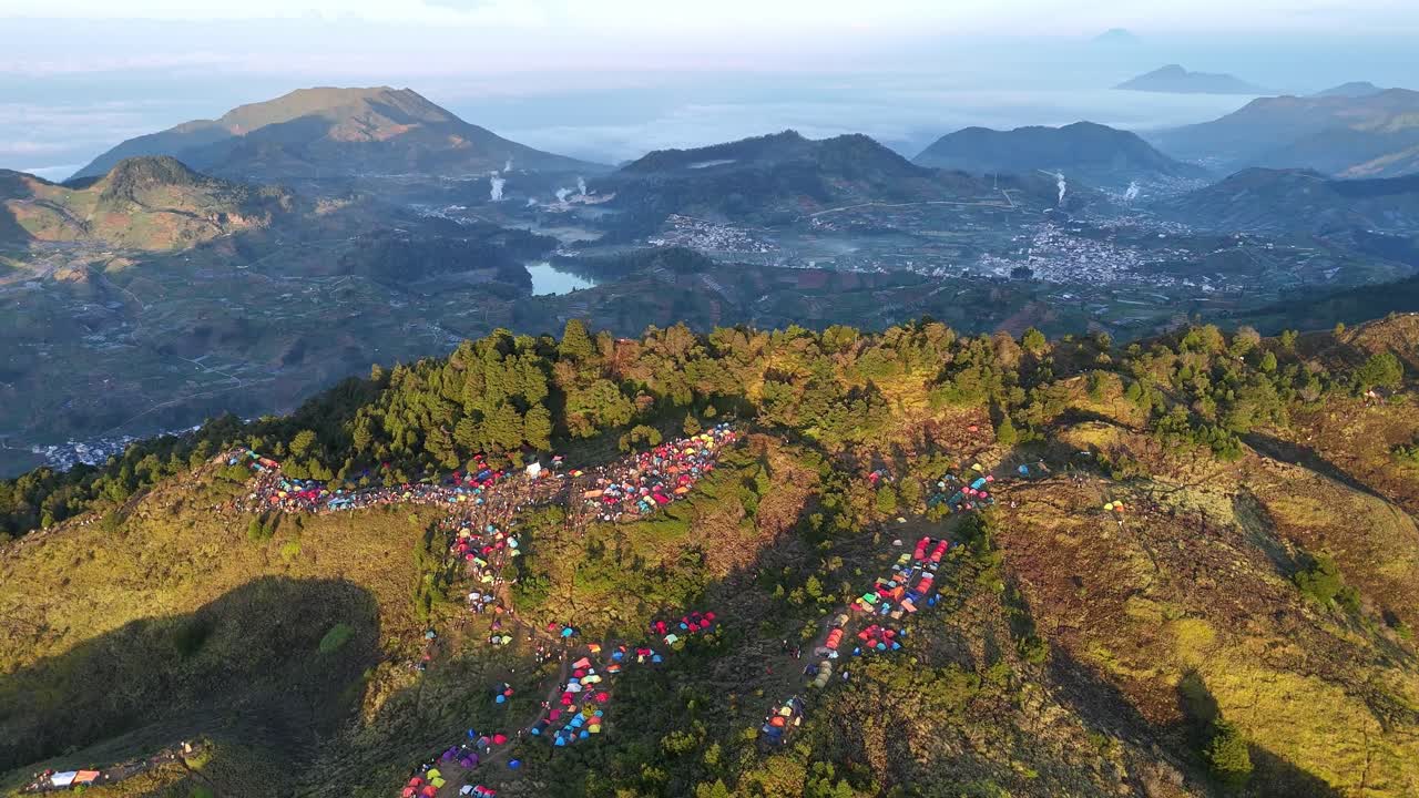 Aerial view of vibrant mountain campsite at sunrise, with rows of colorful tents dotting the hills, dense forest, and a stunning backdrop of lakes, valleys, and distant villages. Mount Prau, Indonesia