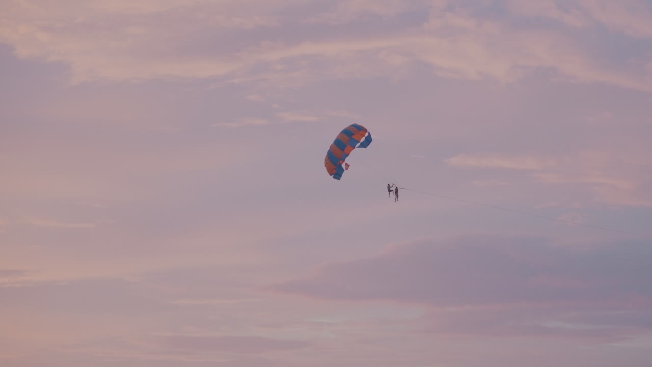 Parasailing al tramonto sopra il cielo aperto dell'oceano, paracadute colorato che sospende un avventuriero solitario nel crepuscolo pastello, deriva calma sopra la silhouette di un'isola lontana, atmosfera di svago serena, filmato di viaggio cinematografico all'ora d'oro.