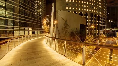 Timelapse-Presenta-Un-Puente-Peatonal-De-Madera-Cálidamente-Iluminado-Con-Barandillas-De-Metal-Por-La-Noche,-Con-Un-Telón-De-Fondo-De-Edificios-Modernos-Y-Brillantes-Con-Arquitectura-Curva