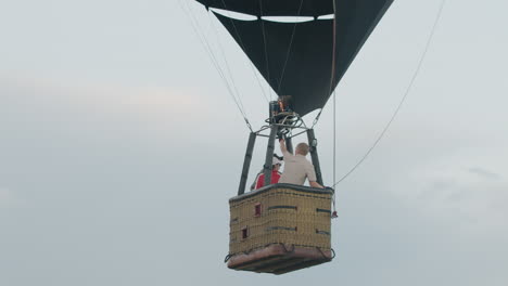 three people inside hot air balloon during pre flight phase as man in white top adjusts flame intensity and checks control device while others observe calmly under early morning sky