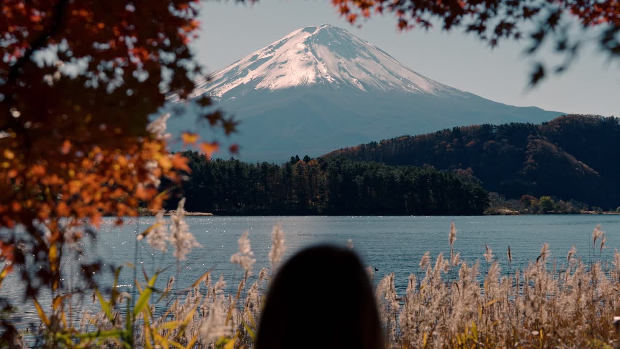 una serena toma de inclinación amplia que muestra la impresionante belleza del monte fuji desde el lago kawaguchiko en otoño y se ve a una mujer mirando el impresionante paisaje.