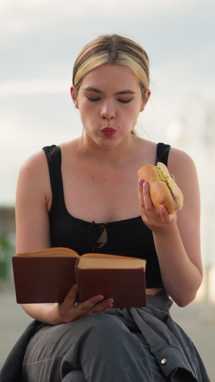 mujer sentada al aire libre con las piernas cruzadas sosteniendo un libro en una mano mientras come un rollo de salchichas y lee, el fondo presenta un poste de lámpara y personas caminando en la distancia
