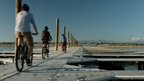 three friends ride their mountain bikes over an empty pier at antelope island state park in utah, the shoreline and mountains are in the distance