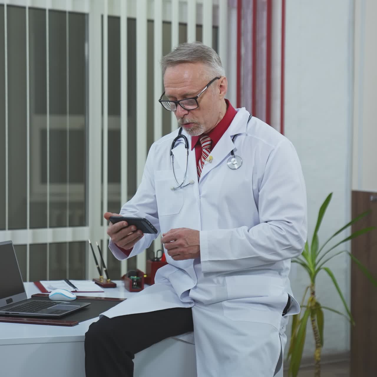 Consulto medico durante la quarantena. Medico anziano con gli occhiali seduto nel suo studio e in videochiamata con un paziente usando il telefono.