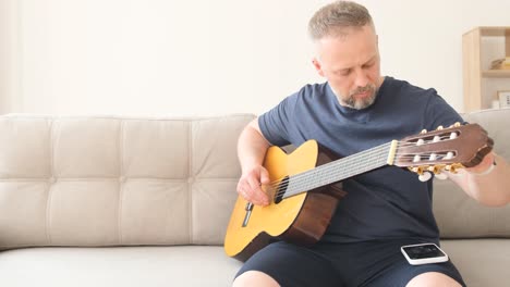 a male musician, guitarist tunes an acoustic guitar. the man twists the tuning pegs on the guitar and listens carefully to how the strings of the acoustic guitar sound. the musician tunes the guitar connected to the laptop
