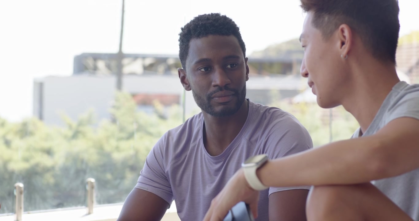 Asian man wearing smartwatch showing smartphone to African American man watching content on balcony