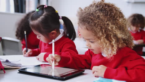 Two-infant-school-kids-sitting-at-a-desk-drawing-with-a-tablet-computer-and-stylus,-close-up