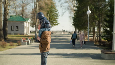 a back view of a father carrying his child on his shoulders while walking down a pathway then turned around, with other people walking ahead and a green-roofed building in the background