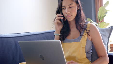 Focused-biracial-teenage-girl-on-couch-using-laptop-and-talking-on-smartphone,-slow-motion