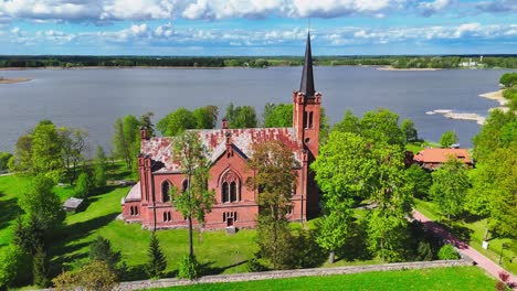 a pointed-steeple church built from red brick rests near a lakeshore, framed by spring trees and dramatic skies in a serene european landscape.