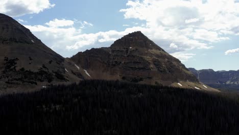 Stunning-aerial-drone-shot-of-a-large-pointy-rocky-mountain-with-snow-caps-and-the-large-Uintah-National-Forest-below-on-a-warm-summer-day-in-Northern-Utah